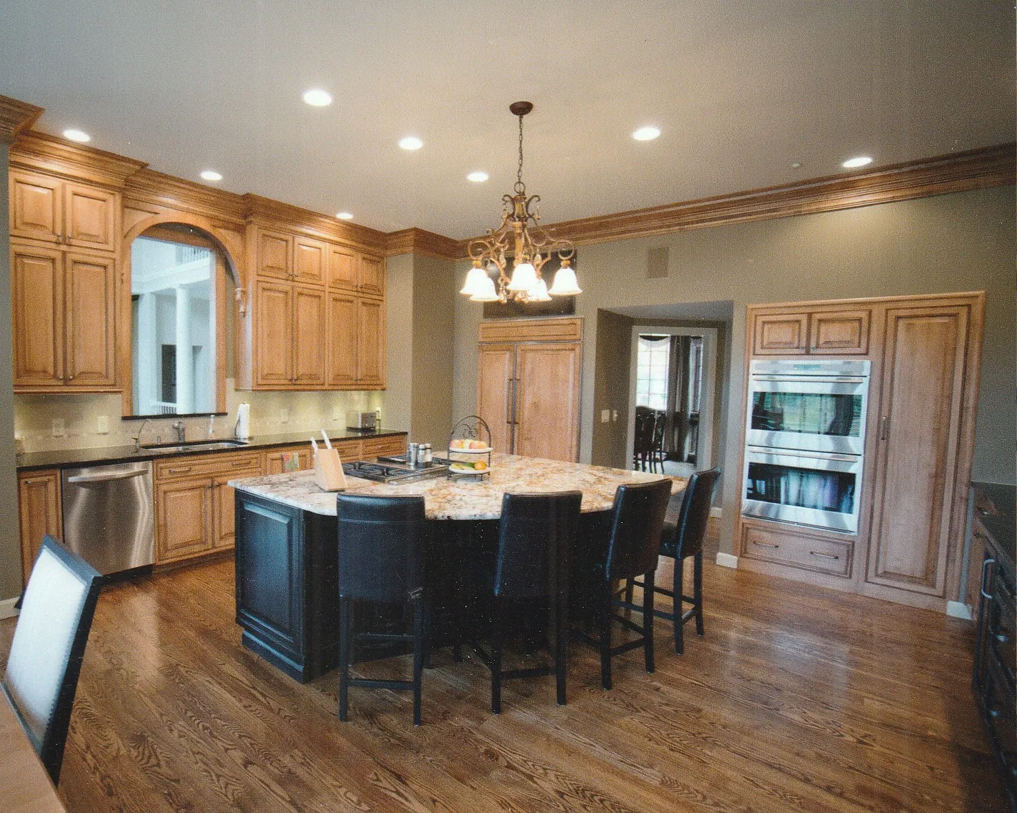 Spacious kitchen with wooden cabinetry, granite island, and modern appliances, featuring a chandelier and warm hardwood flooring